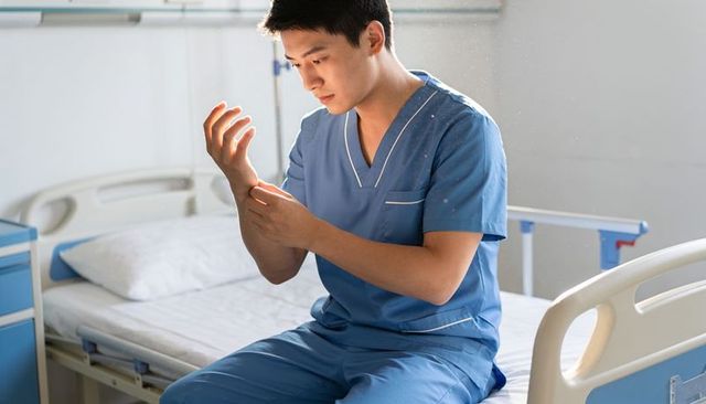 Asian male nurse checking wrist while sitting on hospital bed in blue scrubs