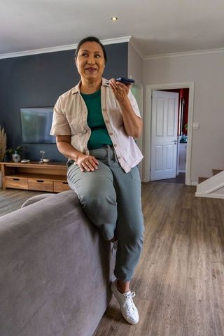 Mature woman sitting on sofa holding smartphone and speaking, relaxed home lifestyle