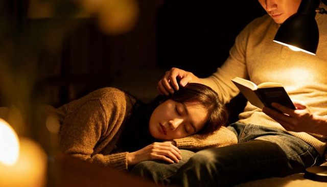 Couple cuddling on sofa with woman resting on lap while man reads under warm lamp glow