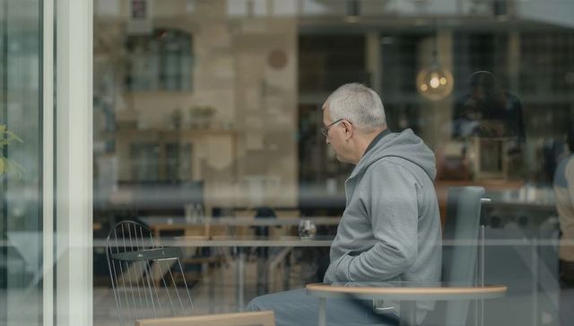 Senior man gazing through cafe window sitting in chair with coffee cup and reflections