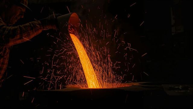 Molten metal pouring into mold with flying sparks, foundry worker guiding ladle