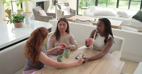 Diverse women sharing coffee and conversation around wooden dining table in bright home