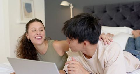 Couple relaxing on bed using laptop, smiling together in cozy modern bedroom