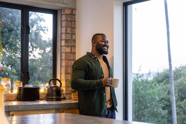 African american man sipping coffee and smiling in modern cozy kitchen by window