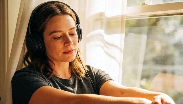 Serene woman listening to music at sunlit window wearing over-ear headphones and gray tee