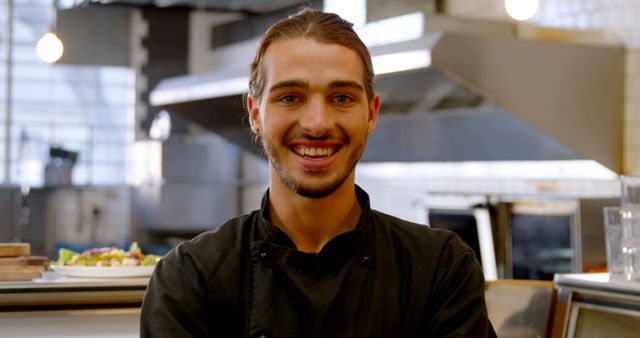 Confident chef wearing black chef coat smiling in modern kitchen environment. Ideal for illustrating culinary expertise, professional chefs at work, restaurant atmosphere, or promotional materials for culinary schools and restaurants.