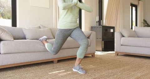 Mid-adult asian woman performing single-leg rear lunge using sofa in sunlit living room