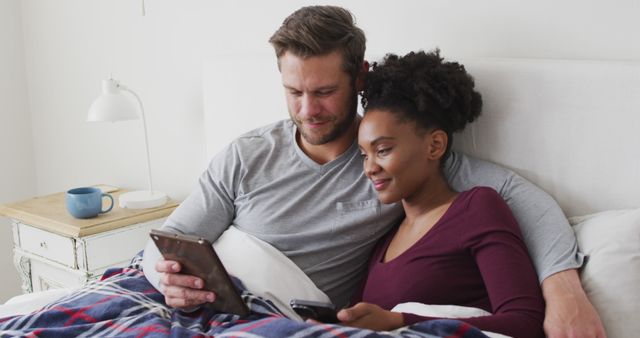 Image of happy diverse couple using tablet in bed and talking. Love, relationship and spending quality time together at home.