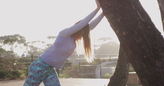 Woman performing a stretching exercise by leaning against a tree in a sunlit park during the morning. Ideal for articles or advertisements focused on fitness routines, healthy outdoor activities, and wellness lifestyles. Can be used to highlight the benefits of starting the day with exercise and enjoying nature.