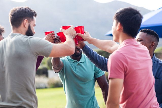 Group of multiracial male friends toasting with red cups at an outdoor weekend party. They are enjoying each other's company against a scenic mountain backdrop. Ideal for use in advertisements, social media posts, or articles about friendship, social gatherings, and outdoor activities.