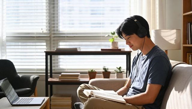 Asian student reading with headphones on sofa at home with laptop and sunlight