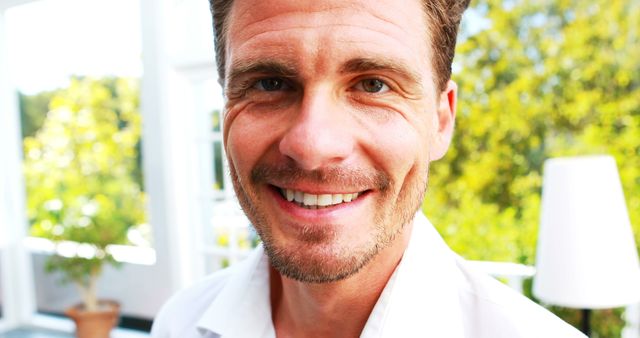 A cheerful Caucasian man smiling warmly at the camera, dressed in a white shirt, outdoors with greenery in the background. The bright sunlight and modern setting provide a vibrant and positive mood, making this image perfect for promoting lifestyle blogs, health and wellness campaigns, advertising casual attire, or portraying relaxation and happiness in marketing materials.