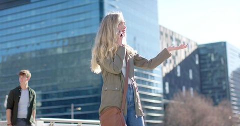 Young woman talking on smartphone while gesturing on sunny urban plaza near glass towers