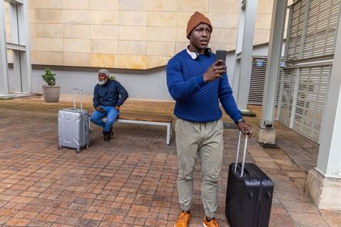 Young african american man checking phone while pulling suitcase in urban courtyard
