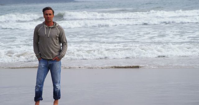 Man standing barefoot on sandy beach while wearing casual outfit with hoodie and jeans, looking at camera. Waves and ocean provide serene backdrop. Ideal for themes of relaxation, travel, leisure, coastal living, and peaceful outdoor moments.