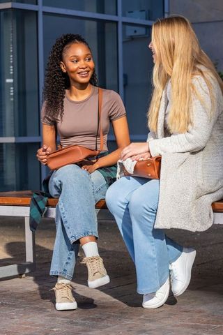 Sunlit diverse friends chatting on urban bench with leather crossbody bags