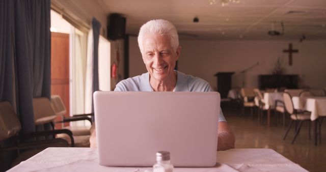 Elderly man with grey hair smiling and engaging with his laptop at a community center. The setting features tables and chairs, utility equipment, and a relaxed atmosphere. This image can be used in articles or advertisements about senior activities, technology use among seniors, lifelong learning, community gatherings, or digital communication tools for the elderly.