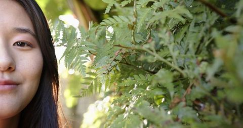 Mid adult asian woman showing left profile beside sunlit ferns, closeup with copy space