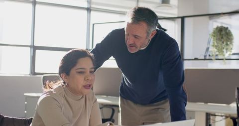 Senior mentor guiding diverse coworker reviewing documents on laptop in bright office