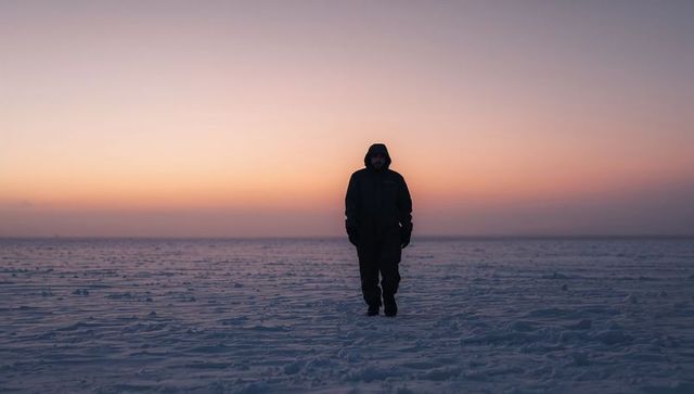 Walking hooded man crossing vast snow plain at dusk, solitary winter silhouette