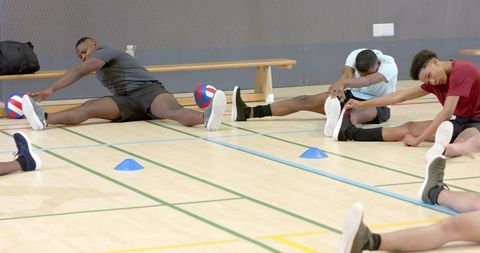 Diverse male teammates stretching and reaching toes during volleyball warmup in gymnasium