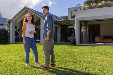 Young couple standing on lush lawn in front of modern home with open patio and pergola