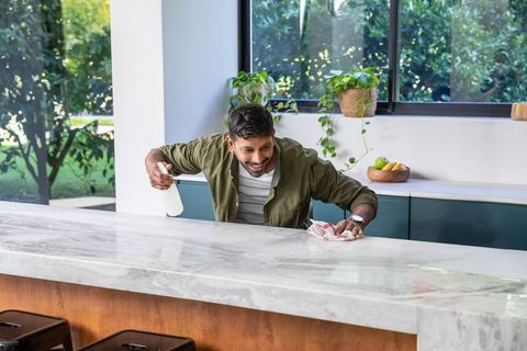 Mature indian man cleaning marble kitchen island, spraying and wiping countertop