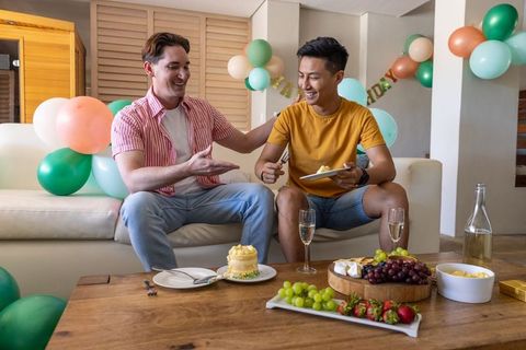 White and asian male friends celebrating birthday on cozy living room sofa with cake and balloons