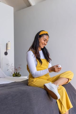 Smiling asian woman sitting on bed wearing yellow overalls and headband holding white case