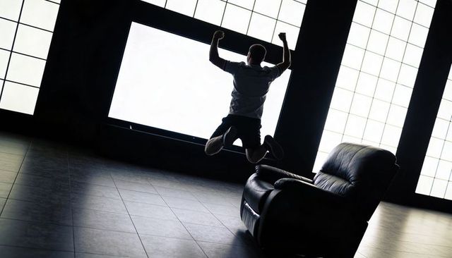 Silhouette man jumping with arms raised in dramatic backlit lounge with recliner