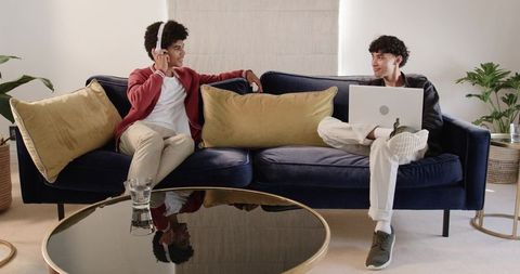 Diverse young friends relaxing on navy sofa sharing headphones and laptop in modern living room