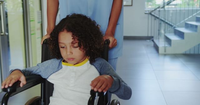 Young child with curly hair sits in wheelchair, pushed by nurse in uniform through hospital corridor. Sad facial expression suggests concern. Image useful for illustrating healthcare services, patient care, pediatric health, or emotional moments in medical settings.
