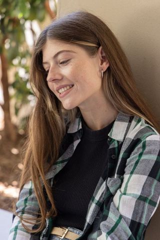 Smiling young woman leaning on stucco wall wearing plaid shirt and small hoop earring