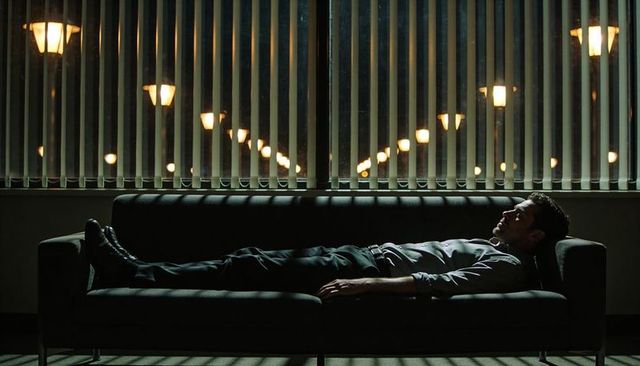Late-night executive reclining on office sofa with blinds casting streetlamp reflections