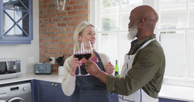 Biracial senior couple wearing aprons toasting wine glasses while cooking in the kitchen at home. retirement senior couple lifestyle living concept