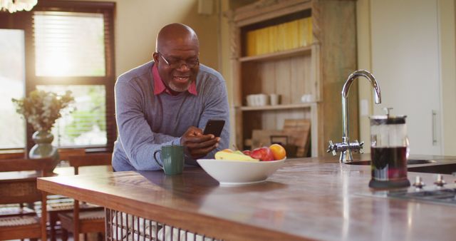 African american senior man leaning on kitchen counter using smartphone, taking off glasses, smiling. retirement lifestyle, spending time alone at home.