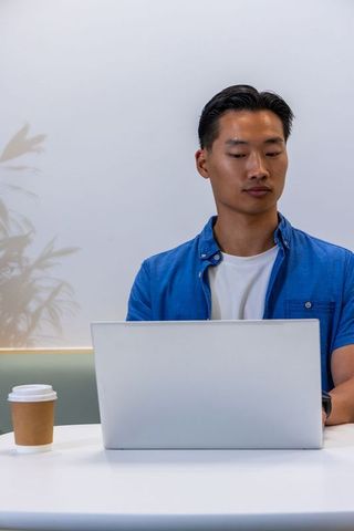 Asian man working on laptop in minimalist cafe workspace with coffee cup and calm focus