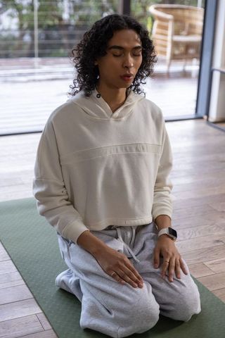 Non-binary person meditating on mat in minimalist home wearing cream hoodie and smartwatch