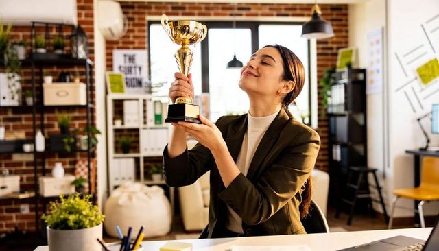 Young woman celebrating holding gold trophy at modern office desk, coworking startup success