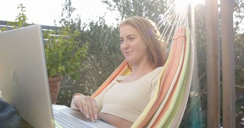 Woman working on laptop in hammock under sunlit pergola