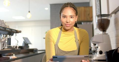 Smiling barista holding tablet at counter wearing yellow top and gray apron