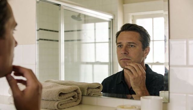 Man grooming and checking chin in bathroom mirror with towels and soft window light