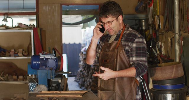 Male carpenter in workshop talking on phone while working on woodworking project. Dressed in apron, surrounded by tools and machinery. Ideal for themes of craftsmanship, artisan skills, DIY projects, small business operations, industrial labor, or professional craftsmanship.