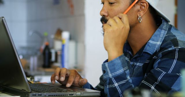 Man sits in a casual kitchen environment working on a laptop while talking on a cell phone. Ideal for themes of remote work, multitasking, technology use, home office setup, and productive work habits.