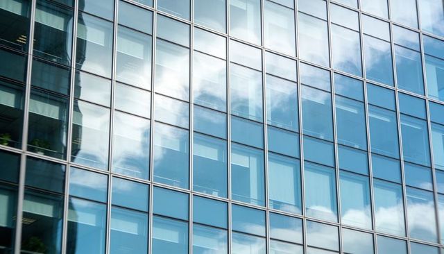 Glass facade mirroring sky and clouds with visible office desks, plants and mullions