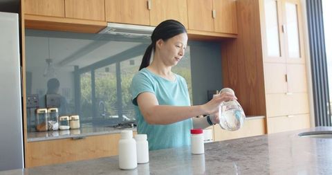Asian woman pouring water into supplement container on minimalist modern kitchen island
