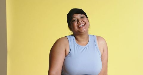 Laughing plus size woman in black cap and blue athletic top on yellow background