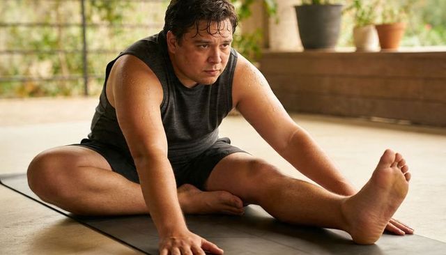 Plus-size man stretching on yoga mat on patio, sweating, focusing on flexibility
