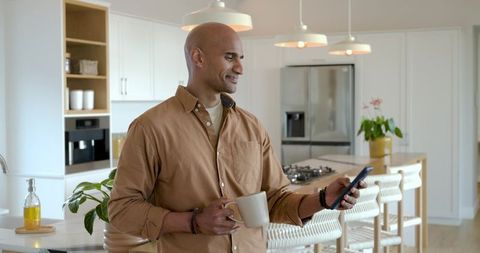 Mature man checking smartphone while holding ceramic mug in bright modern kitchen