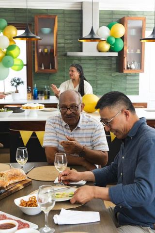 Diverse senior friends celebrating at home kitchen table with food, balloons and laughter